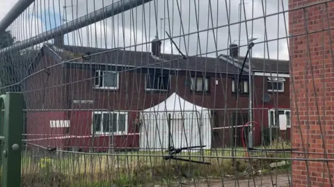 A brown block of flats can be seen through a wire fence. A white police tent, which is surrounded by police tape, has been set up next to the building.