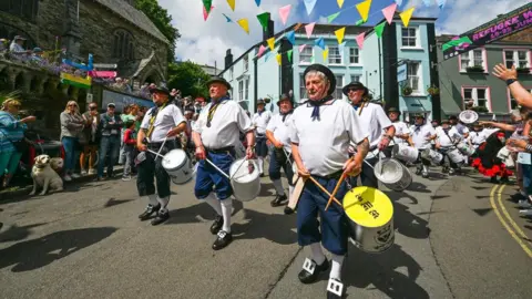 Hugh Hastings A sea shanty band walks down the street. The large group are wearing white shirts and blue shorts. They are all carrying a drum on their left hip. People line the streets to clap them.
