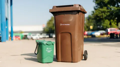 A large brown bin with wheels is situated next to a small food waste green caddy with a lid and handle. They are placed on a street next to a car park which is blurred. It says 'Hull recycles' in white lettering on both of them.