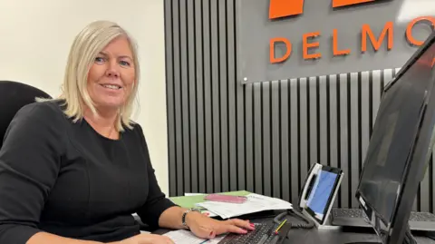 Angela has blonde hair and is wearing a black top. she is sitting at a computer screen.