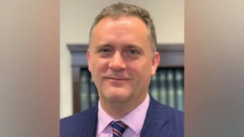 Coroners' Society Lincoln Brookes, who has short grey hair and is smiling at the camera. He is wearing a navy, pinstriped suit jacket with a pink shirt and blue and red tie.