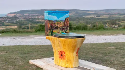A painted yellow toilet on a wooden pallet in a landscape of fields and rolling hills.