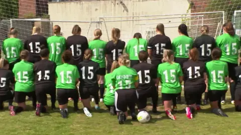 22 people lined up with their backs to a football net. Half are wearing green T shirts and half are wearing black T shirts, they all have the number 15 on them in white letters