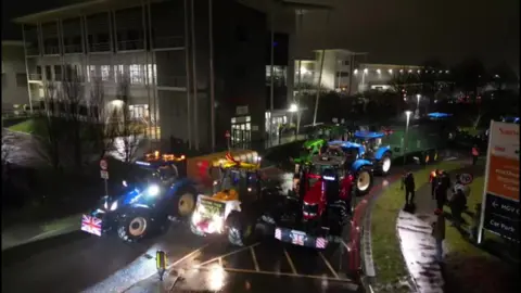 A vantage point view of lots of tractors with their lights on outside a large building. It is still dark.