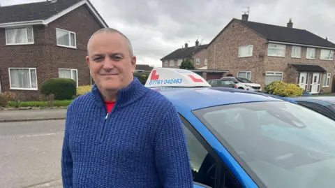 Dave Vasey, a clean shaven man with short grey hair in a blue quarter zip jumper with a red tshirt peaking through is standing next to a blue car with a learner sign on the top. He is in a residential street