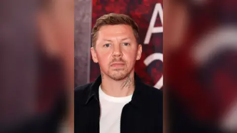 Professor Green has short blonde hair and is wearing a white t-shirt under a black jacket