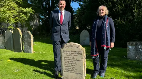 Ben Prater Chris Brooks and Cristina Staff standing smartly dressed in sunshine behind the headstone in the grounds of Malmesbury Abbey. 