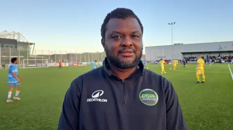 Adekanmi Abayomi, a man with short, slightly greying dark hair and a matching beard, looks at the camera while standing on a football pitch on a sunny day. He is wearing a black half-zip branded sports top with a EMSONI badge. There are players on the pitch behind him. 