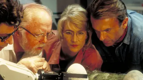 Getty Images From left to right, actors Jeff Goldblum as Dr. Ian Malcolm, Richard Attenborough as John Hammond, Laura Dern as Dr. Ellie Sattler and Sam Neill as Dr. Alan Grant, watching a robotic arm handle the dinosaur eggs in a scene from the film 'Jurassic Park', 1993.