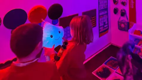 Image of a girl with a short bob hairdo, holding a blue smiling star shaped toy.  Standing next to her is a young boy who is watching. The room is glowing red.  In the background can be seen a  number of games boards on the walls, large round discs and headphones.