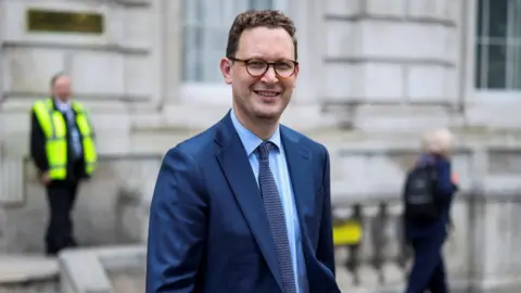 Darren Jones, who has short brown hair and glasses, smiles at the camera as he stands in front of the stone-fronted building of Whitehall. He is wearing a medium blue suit, a blue shirt and a dark patterned tie.