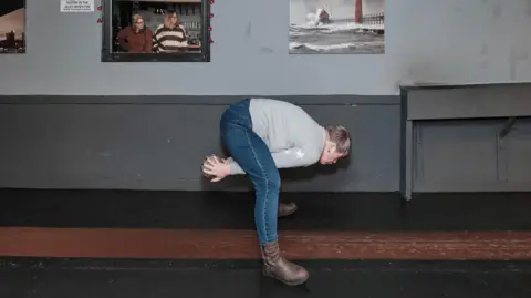 Joss Barratt A woman gets ready to throw a ball down a skittle alley. She is wearing boots, jeans and a grey jumper. There are pictures in frames on a grey-coloured wall behind her.