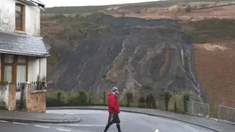 A man in red top and black leggings walks across a road with the damage from an old tip sliding down the mountain in the background