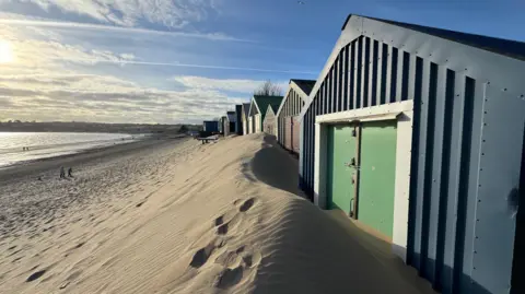 Abersoch.com Rows of sand covered beach huts following storm. People can be seen on the beach and it is a sunny winters day.