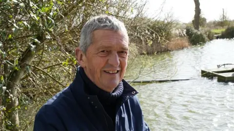 A man smiling at the camera, he is standing next to a reservoir wearing a navy coat with the collar covering his neck. Behind him are some trees that do not have any leaves on them. 