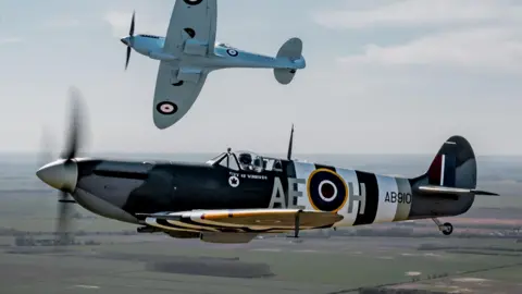 Two Spitfire fighter planes flying over fields under a hazy blue sky. In the foreground, flying horizontally, a Spitfire painted in a green and grey camouflage design with white and black "D-Day" stripes, a large RAF roundel in yellow, blue, white and red and the identification markings AE-H and AB910. In the background, peeling off to the right with the underside of the wings visible, a Spitfire painted in light blue with RAF roundels in blue, white and red.