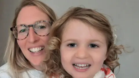 Supplied Megan, with blonde hair, glasses and a grey jumper, posing for a selfie with her daughter Joy, who has curly mousy blonde hair, is smiling and has big blue eyes