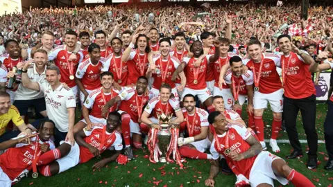 Getty Images The Arsenal men's squad and coaching staff pose for a team photograph on the pitch with the Premier League trophy, surrounded by red and white confetti, with fans visible in the stands behind them.