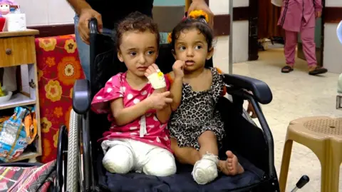 Two sisters are sitting next to each other in a wheelchair, Misk is missing one leg and his sister is missing both legs. Their stumps are bandaged and Hanani raises his hand which is also bandaged