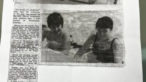 News Quest Black and white newspaper cutting from May 1965 showing two 16-year-old girls in a swimming pool. Both have short dark hair, the girl on the right is Hilary Peck who later became Hilary Butler and is wearing a dark swimsuit, her friend on the left is wearing a white one.