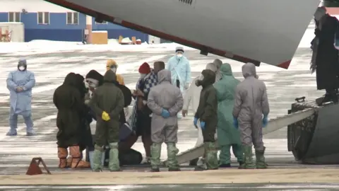 Tyumen regional government Passengers go past specialists in protection suits as they leave an Ilyushin Il-76MD military transport plane which delivered Russian citizens from China at the Roshchino International Airport outside Tyumen, Russia, 05 February 2020