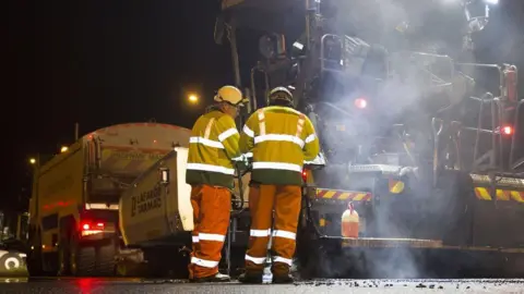 Two workmen stand in front of large machinery with tarmac lettering on it. They are wear bright reflective clothing and the machinery has reflective markings on it. There is steam coming from fresh tarmac laid on the ground. 