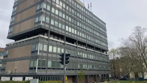 A grey and glass seven-storey office block with traffic lights and some bare trees outside