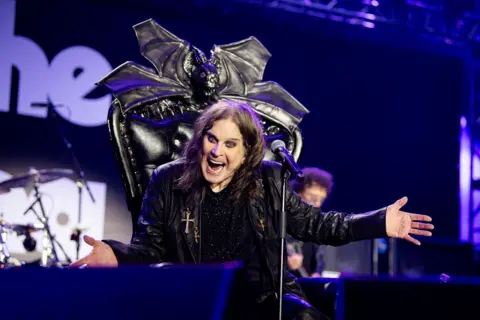 Ozzy Osbourne, who has long brown hair and heavy eye make-up, wears a black leather jacket while sat on a black gothic throne