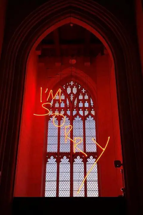 Dominic Mason The words "I'm sorry" in neon yellow lettering hanging down with a large church window in the background.