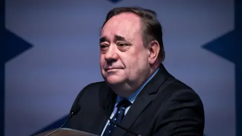 PA Media Alex Salmond, with dark hair and wearing a dark suit, sitting in front of a saltire at a press conference 