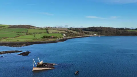 A half sunken ship sticking out of the water in Strangford Lough, there is a small boat sailing near it in the water, in the distance is land.