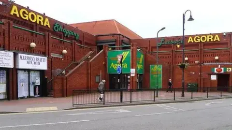 A two-storey red brick building occupying the corner of a site. The word "AGORA" is visible towards the top of the sides of the building and over an entrance in the apex of the corner. The building is divided into shop units, including a post office and a beauty salon. There is a paved pavement area separated from a road by black railings.