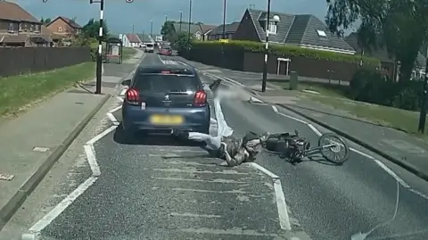 Northumbria Police A still from a video which looks like dashcam footage showing a car on a residential road, and alongside it on the floor is a bike and its rider, who has his legs in the air.