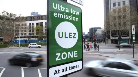 PA Media A sign saying 'ultra low emission zone - at all times' in green and white, at a road junction in near Tower Bridge with cars and buildings in the background.