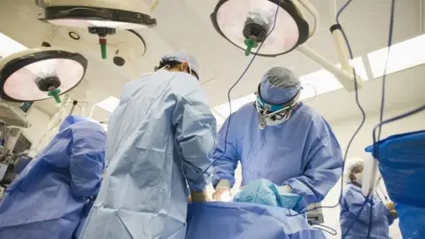 Three surgeons wearing blue gowns and facemasks operate on a patient in a surgical ward 