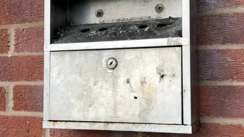 A silver cigarette bin, cover in ash, is mounted on a red brick wall.