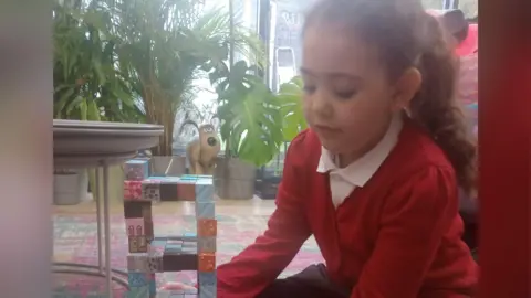 PA Media Ellaria wears a red cardigan and white polo shirt. She is playing with building bricks in a room full of plants behind her. 