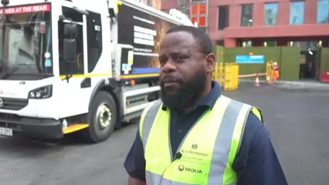 A man wearing a yellow hi-viz Veolia branded jacket, with 'City of Westminster' on it. He has short black hair and a beard. He is stood in front of a rubbish truck. 
