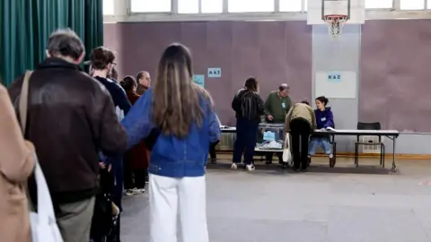 Voters queue inside a polling station at the far end of which two officials at a long table face two of the voters about to retrieve their ballots