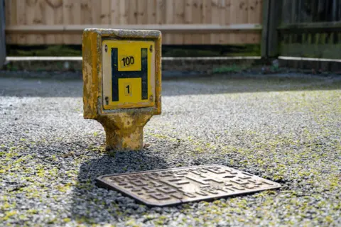 Northamptonshire Fire and Rescue Service Close-up of a yellow fire hydrant marker post labelled ‘H 100 / 1’ next to a metal street drain on a paved surface.