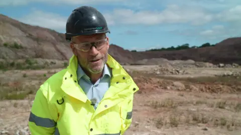 Jeff Court wears a black hard hat, blue shirt, yellow high-viz and a smile as he looks off to the side while standing in front of a rocky area of mining