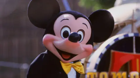 Getty Images The Disney character Mickey Mouse in a Disney World theme park, smiling widely and wearing a gold bow-tie. 