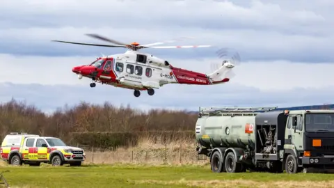 Eshott Airfield A red and white coastguard helicopter comes in to land next to a fuel truck and airport pick up truck