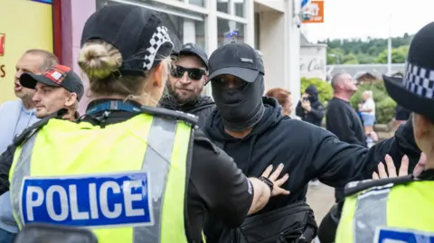 PA Media A police officer with her hand on the chest of an anti-immigration protester wearing all black. His face is covered and he is wearing a baseball cap. There are other people and another officer on the picture.