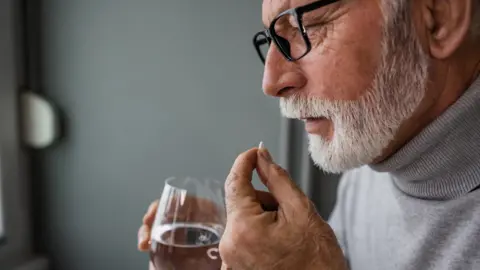 Getty Images A man with a white beard holds a glass of water while taking a pill