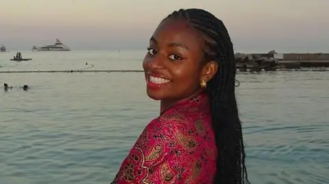 A portrait of Clara Bale smiling, with braided hair and wearing a patterned pink garment, standing near the water at dusk.