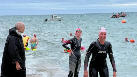 A group of people at a sea slip, some wearing wetsuits and swim caps, with several swimmers in the water and a few boats in the background under a cloudy sky with visible sea waves