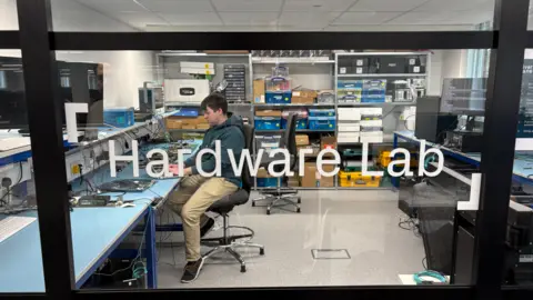 Ben Schofield/BBC Looking through a window with 'hardware lab' written in white text on the glass. Inside the room beyond the glass is a man sitting on an office chair working at a computer. His workbench is on the left of the image. There is an empty office chair behind him. There is equipment on the workbench around the laptop and on shelves at the back of the lab. 