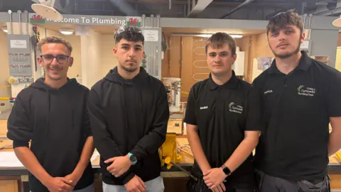 Four young men standing next to each other in a workshop with two on the left wearing black hoodies and the other two wearing black polo shirts and a sign saying 'Welcome to Plumbing' on the wall behind them.