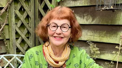 Contributed Marcia Riddington with short red hair and black glasses smiles at the camera while sitting in a garden in front of a shed. She is wearing a green top with a yellow and white striped scarf around her neck.